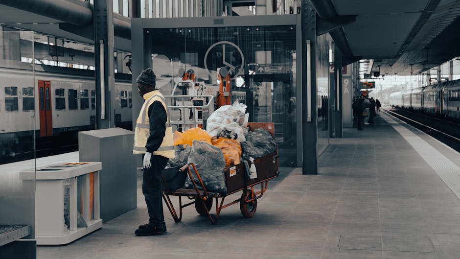 A metal waste disposal bin situated on a train station platform with four color-coded compartments for different types of waste. The left compartment, marked with a yellow label, is designated for pla