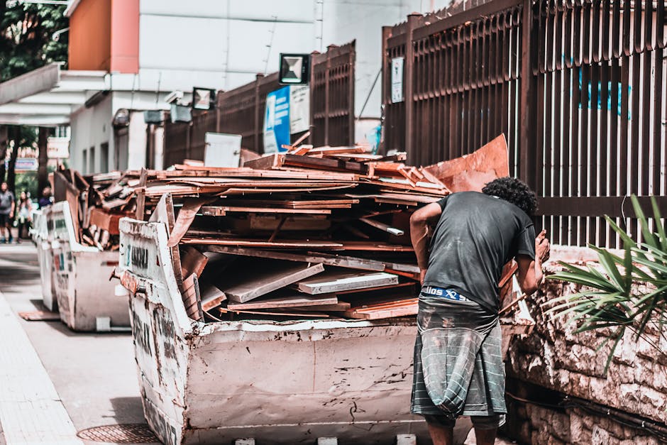 A white van parked on a city street with rear doors open, revealing an interior filled with large black and white plastic rubbish bags, flattened cardboard boxes, and miscellaneous waste materials. Se