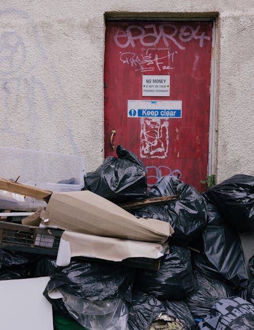 A red metal door set into a beige, textured concrete wall, with graffiti tags in white and pink spray paint on the upper part of the door, and signs indicating 'No Money' and 'Keep Clear' affixed to t